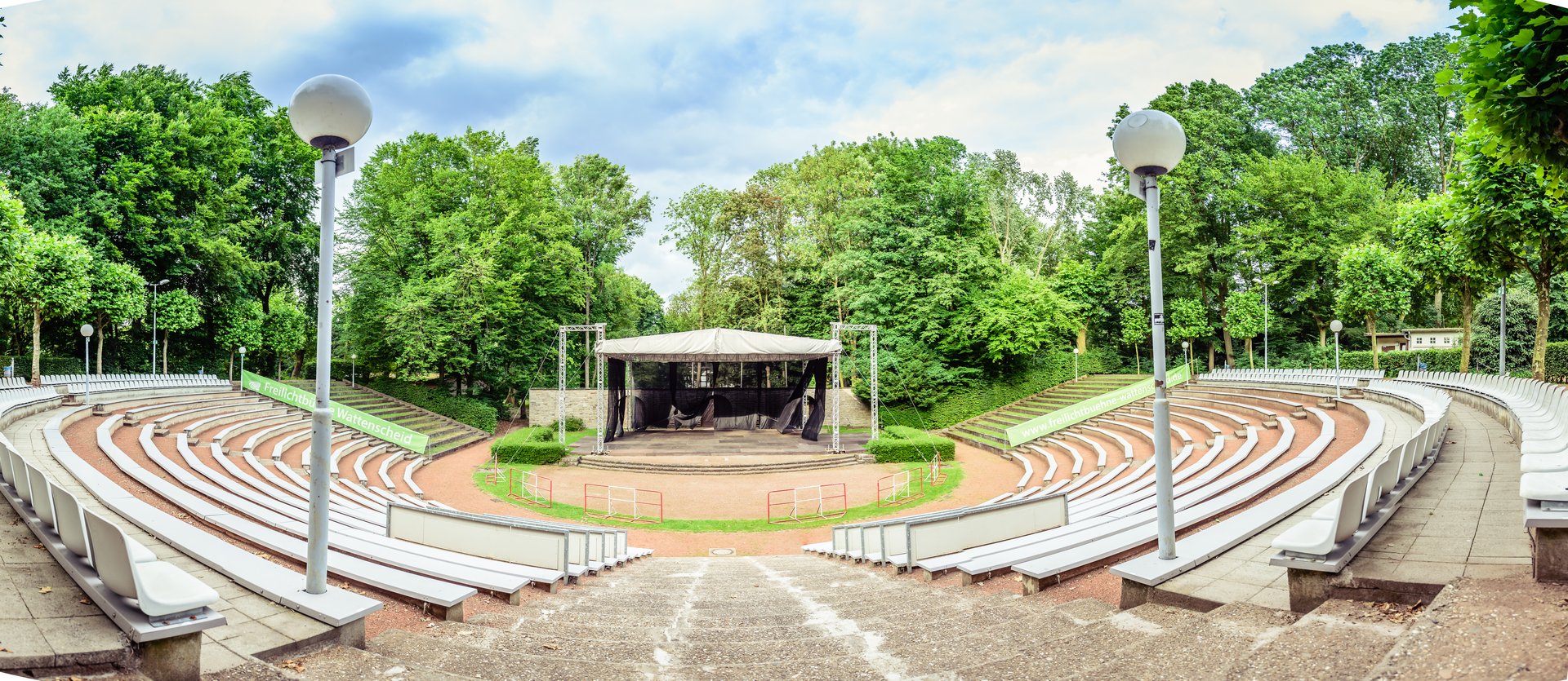 Panorama-Aufnahme der leeren Freilichtbühne Wattenscheid. Im Vordergrund zwei Laternen, die Sitzreihen und die Freitreppe zur Bühne. Im Hintergrund grüne Bäume.