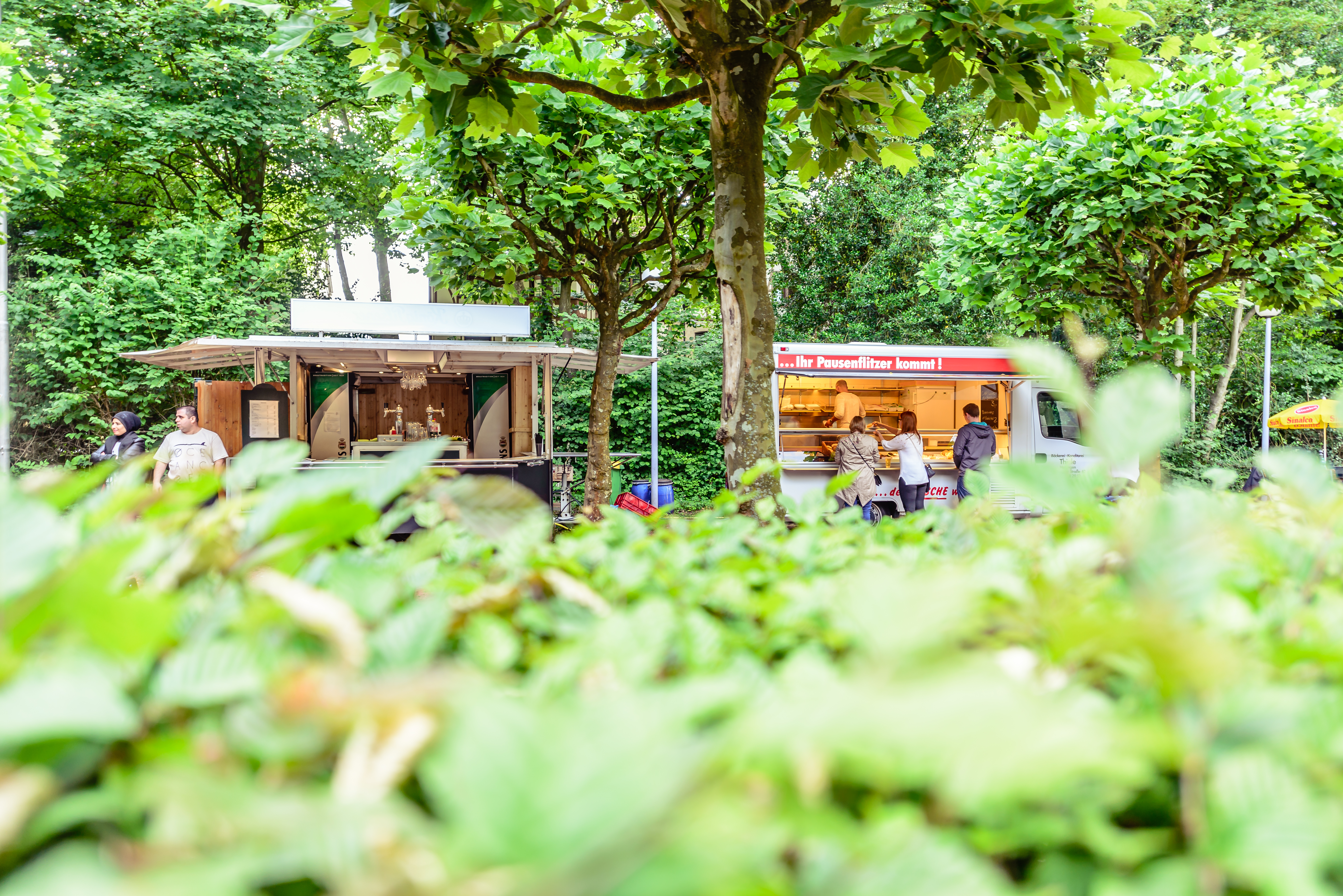 View of two food trucks in the beer garden of the Freilichtbühne Wattenscheid. Green bushes in the foreground.